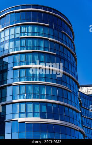 Uno splendido esempio di architettura moderna caratterizzata da una facciata in vetro curvo con elementi visibili in cemento e metallo, adagiati su un cielo blu vivace. Foto Stock