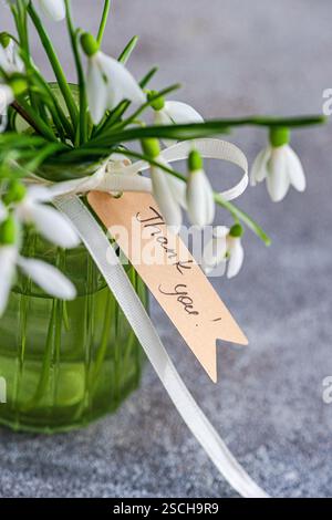 Vista ravvicinata di scintillanti gocce di neve elegantemente disposte in un vaso di vetro verde con un nastro color crema e un cartello di ringraziamento, su uno sfondo grigio sfocato Foto Stock