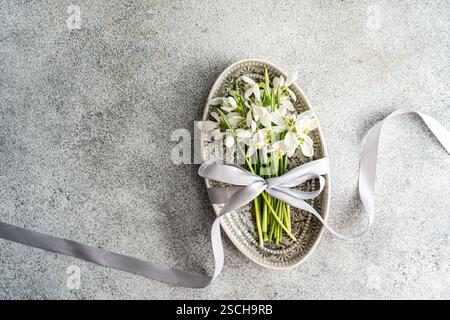 Vista dall'alto delle delicate gocce di neve primaverili legate da un nastro argentato in un piatto vintage decorato su uno sfondo grigio testurizzato. Foto Stock