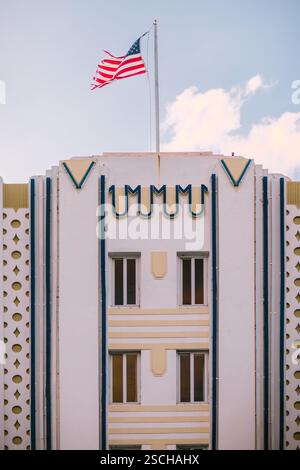 Una vibrante bandiera americana ondeggia sopra un edificio Art Deco color pastello sotto un cielo azzurro, catturando il distintivo stile architettonico e lo spirito di mia Foto Stock