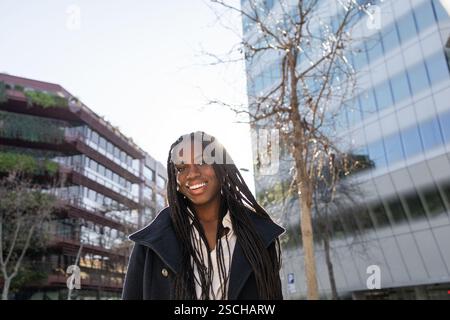 Dal basso, donna d'affari afroamericana sorridente con trecce che si gode una pausa all'esterno dei moderni edifici di uffici, mostrando positività e fiducia. Foto Stock