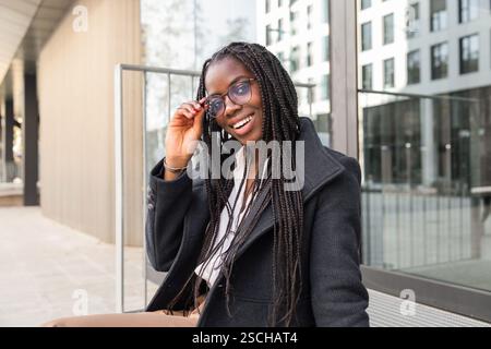 Una giovane donna d'affari afroamericana con i capelli intrecciati si gode una pausa all'aperto, sorridendo mentre si regola gli occhiali. Foto Stock