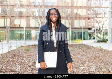 Una donna d'affari afroamericana sorridente, vestita con un elegante cappotto e occhiali, tiene in mano un computer portatile mentre ti godi una pausa all'aperto con edifici moderni all'interno Foto Stock