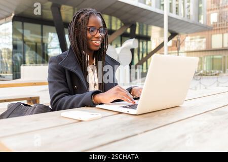 Una giovane donna d'affari afroamericana con trecce lavora su un computer portatile a un tavolo all'aperto in un ambiente informale. Foto Stock