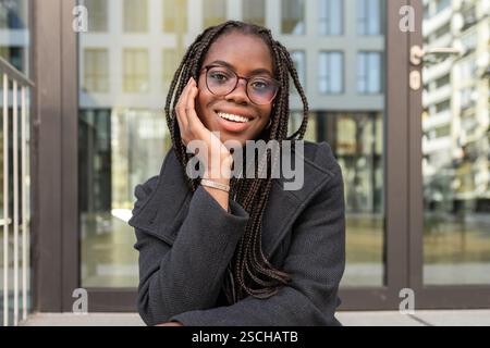 Giovane donna d'affari afroamericana sorridente con trecce, vestita con un abito professionale, che fa una pausa all'aperto mentre è seduta comodamente. Foto Stock