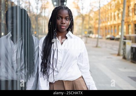 Una donna d'affari afroamericana con trecce è rappresentata all'aperto, indossando una camicia bianca formale e pantaloni cachi, con un espresso focalizzato Foto Stock