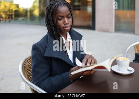 Una donna d'affari afroamericana con trecce è seduta su un tavolo da caffè all'aperto durante una pausa, a leggere un libro mentre si siede accanto a una tazza di caffè. Foto Stock
