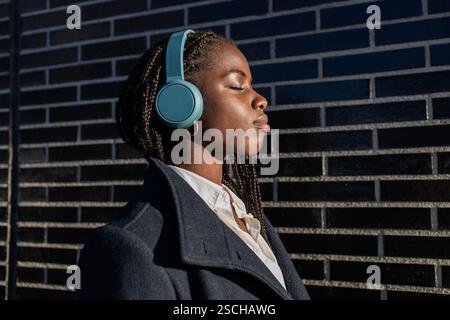 Una donna d'affari afroamericana con trecce si gode una pausa, ascoltando musica attraverso le cuffie, gli occhi chiusi in relax, appoggiandosi a una brilla Foto Stock