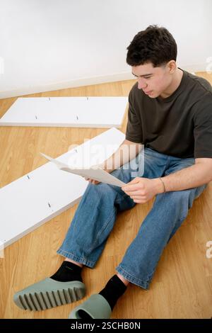 Un giovane uomo siede su un pavimento di legno, leggendo le istruzioni di montaggio per le nuove parti di mobili sparse intorno a lui. Foto Stock
