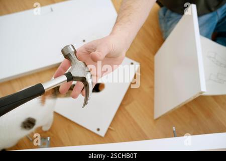 Vista ritagliata di un uomo che tiene in mano un martello mentre assembla nuovi mobili. L'immagine mostra le mani, lo strumento e le parti dei mobili su un flo di legno Foto Stock