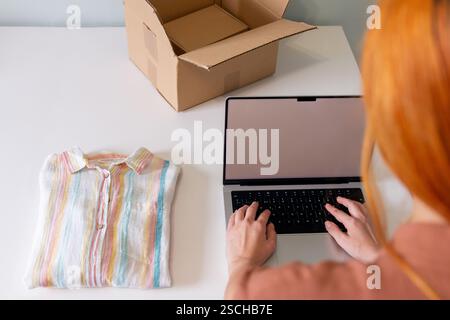Una giovane donna che organizza abiti in vendita, utilizzando un computer portatile, con una camicia piegata e una scatola aperta nelle vicinanze. La vivace camicia contrasta con la scrivania bianca, e. Foto Stock