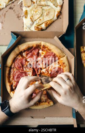 Dall'alto tagliato a mano irriconoscibile che prende fette di pizze assortite da scatole di cartone, mettendo in evidenza uno scenario informale di ristorazione sociale al chiuso Foto Stock