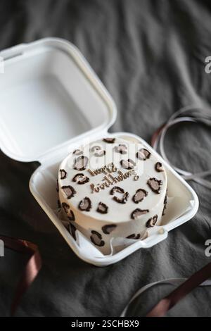 Elegante torta di compleanno bento con stampa leopardata e scritta dorata, ordinatamente presentata in una scatola da asporto bianca su sfondo grigio testurizzato. Foto Stock
