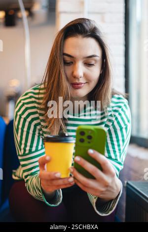 Donna con una camicia a righe che beve un caffè mentre sfoglia il suo smartphone in un bar. Foto Stock