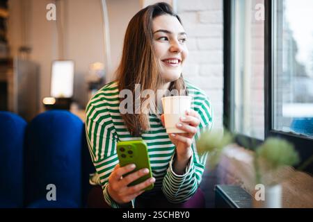 Una donna con una camicia a righe sorseggia un caffè mentre utilizza uno smartphone alla vetrina di un bar, mostrando una pausa rilassata e tecnologica. Foto Stock