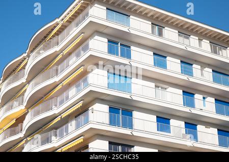 Un moderno edificio di appartamenti presenta diversi balconi curvi, facciata bianca e ringhiere di vetro, adagiate su un cielo blu limpido, che mostra archi urbani Foto Stock