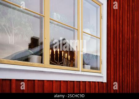 Una calda scena di finestre in Lapponia svedese. La finestra con cornici gialle riflette un intimo interno illuminato da candele con piante in vaso, adagiato contro l'iconico legno rosso Foto Stock