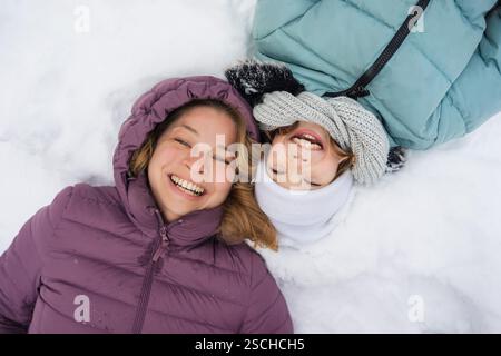 Madre e figlia si godono una giornata invernale giocosa, sdraiati sulla neve. Entrambi indossano giacche invernali colorate, creando un'atmosfera calda e gioiosa in mezzo al c Foto Stock