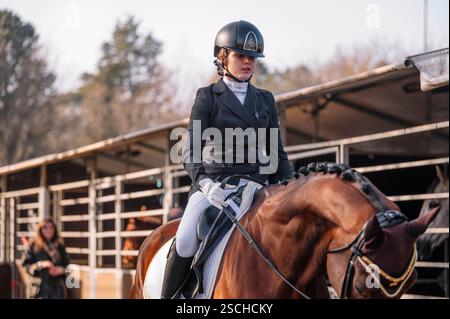 Un'equitazione in abbigliamento completo, casco e guanti che cavalcano un cavallo da baia in una stalla, l'ambientazione cattura l'eleganza e la precisione del classico Foto Stock