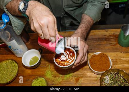 Un barista crea un'intricata latte art su un cappuccino cremoso, circondato da ingredienti come panna, pistacchio e spezie, mettendo in risalto la maestria artistica Foto Stock