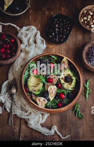 Un'insalata vivace con fichi, avocado, ciliegie, rucola e more in una ciotola di legno. La scena è completata da varie noci e frutta su Foto Stock
