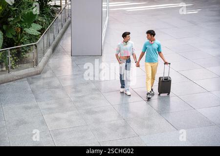 Una felice coppia gay cammina mano nella mano attraverso un terminal dell'aeroporto con i bagagli, godendosi la compagnia reciproca e cercando un abbigliamento elegante e informale. Foto Stock
