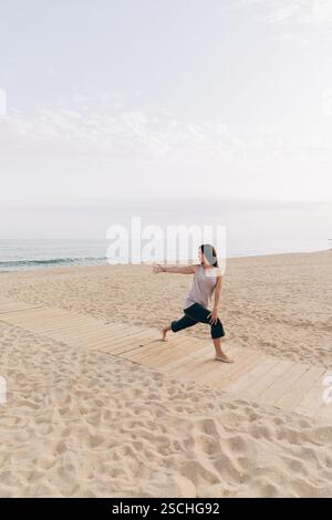 Una donna pratica yoga in spiaggia, mentre posa thea yoga su una passerella di legno su un tranquillo sfondo marino. Foto Stock