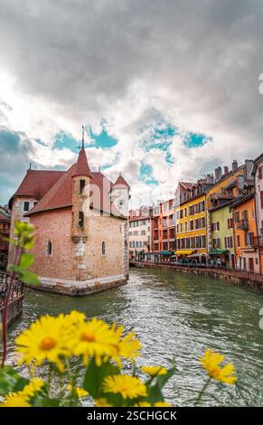 Una vista vivace del Palais de l'Isle e degli edifici colorati adiacenti, adagiati su un cielo spettacolare ad Annecy, in Francia, con freschi fiori gialli Foto Stock