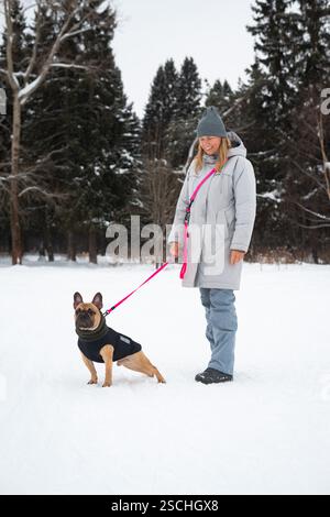 Una persona cammina un cane al guinzaglio in una foresta innevata, entrambi vestiti caldi per l'inverno. La scena serena cattura l'essenza delle avventure in climi freddi Foto Stock