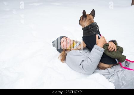 Una persona con un cappotto invernale grigio e un berretto che gioca con gioia sulla neve con un cane vestito. L'ambiente innevato cattura l'essenza del divertimento invernale e. Foto Stock