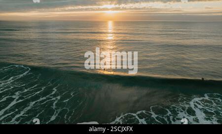 L'immagine cattura un tramonto mozzafiato sull'oceano, con il sole che tramonta in lontananza. Due persone fanno surf sulle onde, godendosi la bellezza Foto Stock