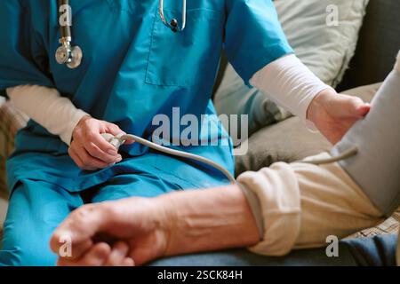 Primo piano medio di infermiera irriconoscibile vestita con scrub e indossa uno statoscopio che misura la pressione sanguigna di un anziano Foto Stock