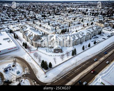 Strada nevosa della città con un grande complesso di appartamenti sullo sfondo. La strada è costeggiata da alberi e la neve è accumulata sul terreno Foto Stock
