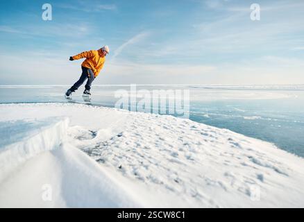 Atleta uomo in giacca gialla pattinaggio su ghiaccio sul lago ghiacciato con crepe e neve in inverno Foto Stock