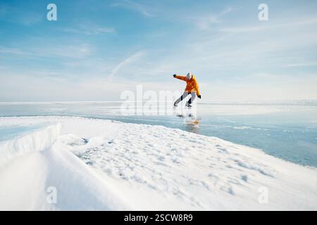 Atleta uomo in giacca gialla pattinaggio su ghiaccio sul lago ghiacciato con crepe e neve in inverno Foto Stock