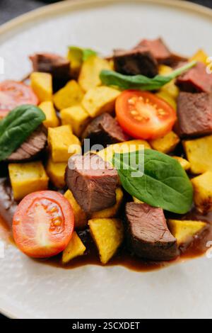 Cubetti di manzo gourmet con patate arrosto, pomodori ciliegini e spinaci con salsa ricca e vino Foto Stock