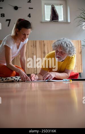 Una giovane donna e un uomo anziano lavorano insieme su un progetto, entrambi impegnati e sorridenti mentre scrivono su carta, seduti sul pavimento in un ambiente accogliente e ben illuminato Foto Stock
