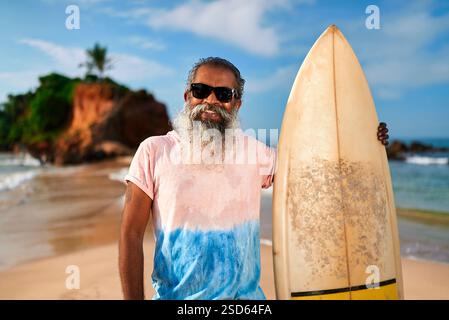 Uomo barbuto anziano con tavola da surf sulla spiaggia soleggiata. Gli anziani surfisti amano uno stile di vita attivo vicino all'oceano. Un uomo più anziano con occhiali da sole abbraccia le avventure marine Foto Stock