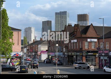 Moulton Street, Manchester, Regno Unito. Traffico su una strada fiancheggiata da negozi, con alti edifici moderni sullo sfondo, tra cui una costruzione Foto Stock