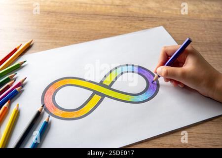 Una mano che incolla il simbolo dell'infinito arcobaleno su un foglio di carta bianco con matite colorate sparse intorno. Concetto di consapevolezza dell'autismo. Foto Stock