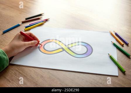 Una mano che incolla il simbolo dell'infinito arcobaleno su un foglio di carta bianco con matite colorate sparse intorno. Concetto di consapevolezza dell'autismo. Foto Stock