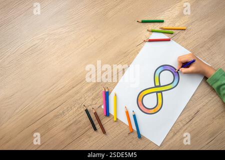 Una mano che incolla il simbolo dell'infinito arcobaleno su un foglio di carta bianco con matite colorate sparse intorno. Concetto di consapevolezza dell'autismo. Foto Stock