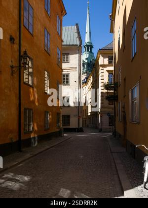 Stretta strada acciottolata nella storica città vecchia (Gamla Stan) di Stoccolma fiancheggiata da alti edifici antichi dipinti - guglia della chiesa tedesca visibile Foto Stock