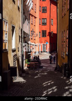 Stretta strada acciottolata nella città vecchia (Gamla Stan) di Stoccolma, Svezia, con edifici dai colori vivaci. La gente si riunisce all'ombra. Foto Stock
