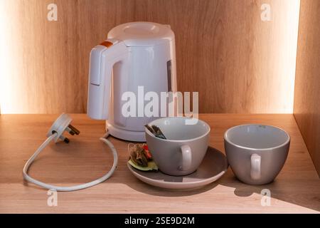 Un bollitore elettrico bianco con due tazze di ceramica, un piattino, bustine di tè e un cucchiaio posto su una superficie di legno, Park Regis hotel, Singapore Foto Stock