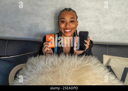 Giovane donna che mostra carta di credito e smartphone seduti comodamente sul divano di casa, shopping online e finanza Foto Stock