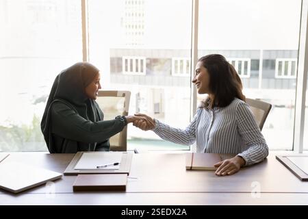Due donne d'affari multiculturali sedute in ufficio sorridenti e stringono la mano Foto Stock