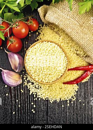 Couscous crudo in una ciotola e un sacco di burlap, aglio, pomodori, peperoni caldi ed erbe su sfondo di legno dall'alto Foto Stock