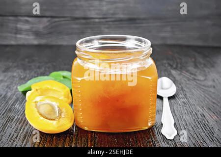 Marmellata di albicocca in vaso di vetro, frutta, un cucchiaio e un ramo con foglie verdi sullo sfondo di legno asse Foto Stock