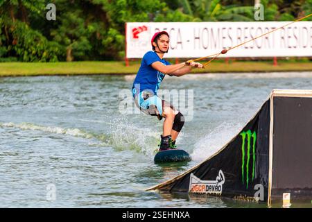 Un giovane adulto maschio wakeboarding a tutta velocità in un parco di wakeboard a Davao City, Filippine, catturato a metà azione Foto Stock